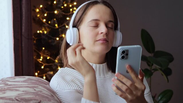 Calm enjoying relaxed woman wearing headphones listening to music online being happy and pleased in Christmas atmosphere sitting on sofa near New Year tree.