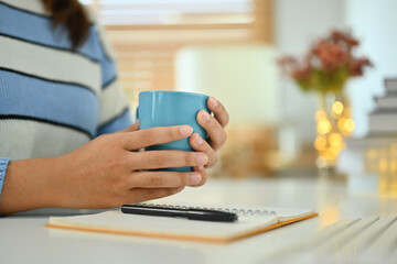 Young woman in warn sweater hands holding cup of herbal tea sitting at workplace