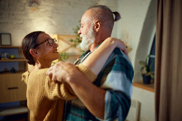 Side view portrait of senior couple, grandfather and grandmother dancing in living room at home and looking to each other with love.