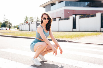 Young beautiful smiling female in trendy summer blue cycling shorts and tank top clothes. Carefree woman posing in street. Positive model having fun. Cheerful and happy. Sits on asphalt. In sunglasses