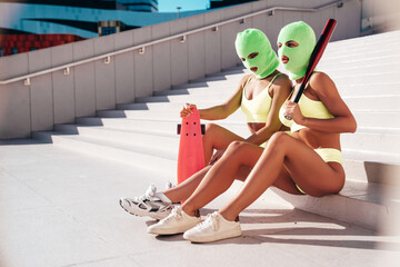 Two beautiful sexy women in green underwear. Models wearing bandit balaclava mask. Hot seductive female in lingerie in street at sunny day. Crime and violence. Hold baseball bat, penny skateboard