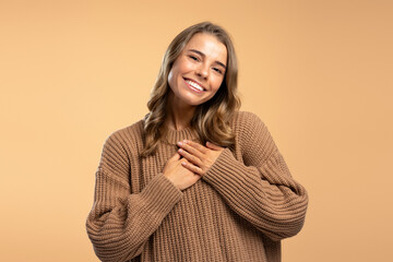 Attractive smiling woman wearing brown cozy sweater holding hands over heart, looking at camera