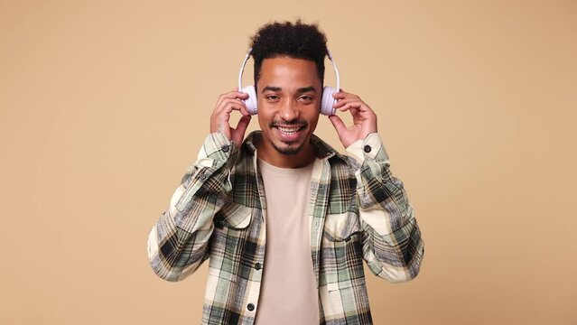 Young man of African American ethnicity he wears shirt casual clothes listen mp3 music in headphones slow dance sing song have fun enjoy relax show thumb up isolated on plain beige brown background