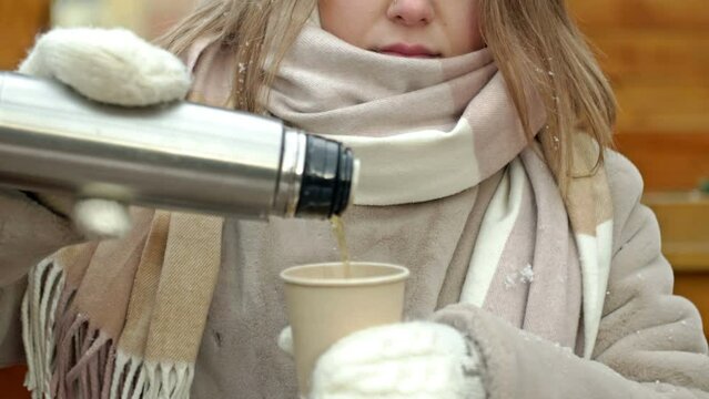 Winter portrait of a young woman in warm clothes pouring herself a hot drink from a thermos. Wind and fine snow.