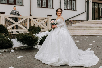 The bride in a white voluminous dress with a long train, holds a bouquet in her hands and poses against the background of the building. Winter wedding.