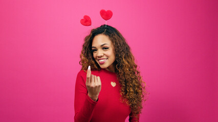 Flirty young woman beckons over secret admirer on Valentines Day, pink studio
