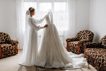 Fototapeta premium A bride in a silk suit and veil poses in her room next to her wedding dress. Morning of the bride. Preparation. Wedding photography