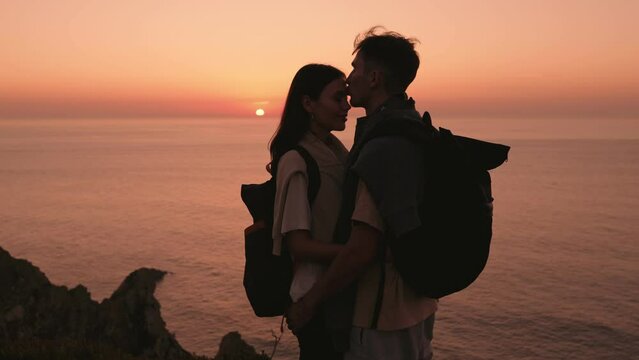 Silhouette of a young couple of travelers in love, sharing a kiss against the peachy sunset over the ocean, symbolizing youth, love, and a deep connection with nature