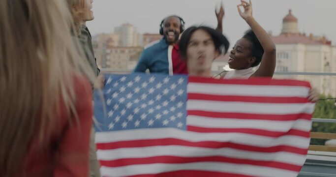 Group of young people dancing with American flag having fun at rooftop party celebrating national holiday. Patriots and modern lifestyle concept.