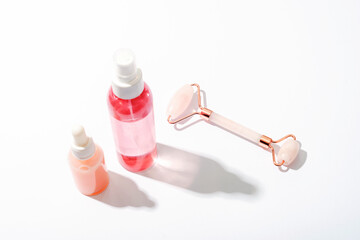 Pink cosmetic bottles and rose quartz crystal facial roller on white background with strong shadows. Natural cosmetics, skin care concept. Top view