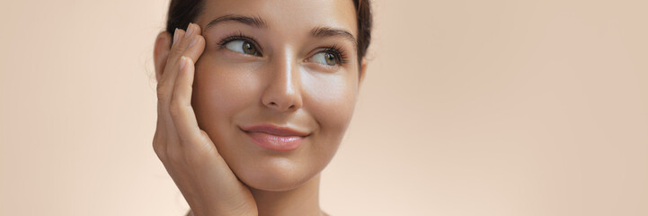 Skincare and Beauty Portrait. Woman with beautiful face touching healthy facial skin. Young adult smiling Asian girl model with natural makeup touching glowing hydrated skin on beige background