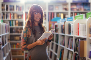 Young smiling Caucasian woman with tattoo holds book and looking at camera posing in bookstore....