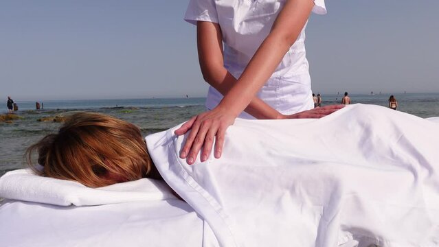 Woman having relax in tropical massage spa on the beach near the sea on massage table. Professional masseur provides back and neck spa procedures. 