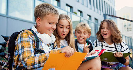 Group of elementary school children writing in their copybooks having an outdoor classwhile sitting on stairs of modern school building background.