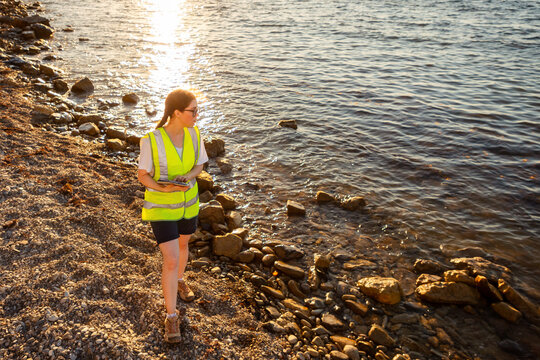Top view of young volunteer woman wearing vest and using pad for analyzing. In background is ocean, coast and sunset. Copy space. Concept of inspection environmentalist and ocean pollution