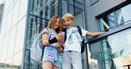 Two teenage students boy and girl friends using smartphone exchange lectures near school building summer day on stairs outside campus. Friendship. Back to school.