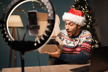 Male influencer wearing Santa hat recording a video with his smartphone in a ring of light while...