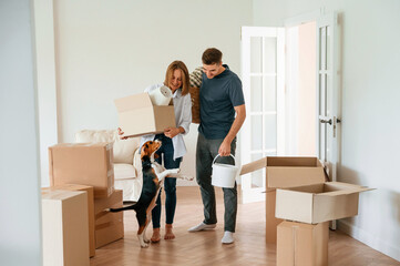 In the big room, with boxes in hands. Young couple with dog are moving to new home - obrazy, fototapety, plakaty