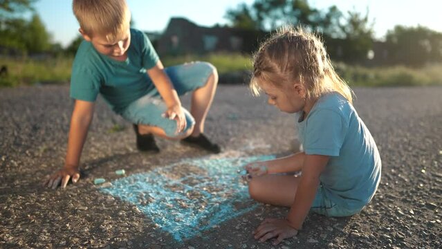 girl and boy are drawing house on the asphalt. happy family kid concept. girl boy sit on the asphalt during the day and draw home big with dream blue chalk. girl boy draw with chalk on the asphalt