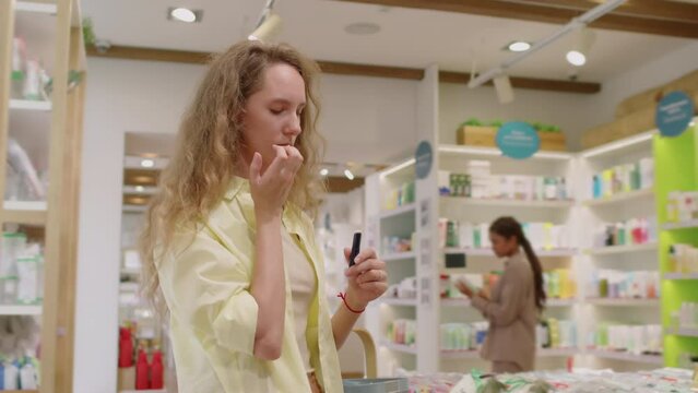 Medium shot of female client choosing lipstick during shopping in cosmetics store