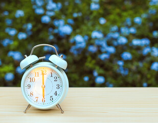 white vintage alarm clock showing 6 o clock on the table in the garden with purple flowers background.