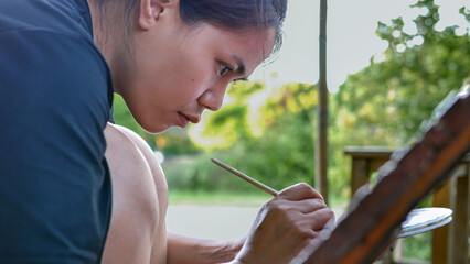 Close-up of Asian woman painter creating art use a paintbrush to draw lettering designs on a wooden coffee shop sign.outdoor activities,People doing activities. - obrazy, fototapety, plakaty