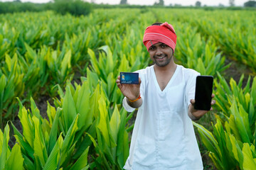 Young indian farmer showing debit or credit card at his green agriculture field, using mobile phone