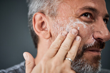 Fototapeta premium Close-up of a mature man, applying a face cream on his cheek, using his fingers.