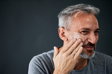 Middle-aged gray-haired man putting face cream on his cheek.