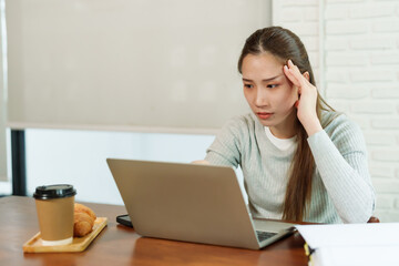 Beautiful Asian business woman in gray sweater sitting  working in front of laptop, looking stressed, holding hands on emples. Next to it croissant brown disposable coffee cup. Insmall business cafe