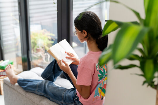 Girl sitting at hassock near window and reading book