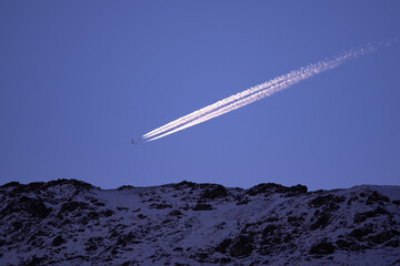 An aircraft flying in the blue sky very close to the mountain peaks. Passenger plane emitting smoke...