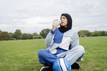 Portrait of woman in hijab sitting in soccer field, drinking water from bottle