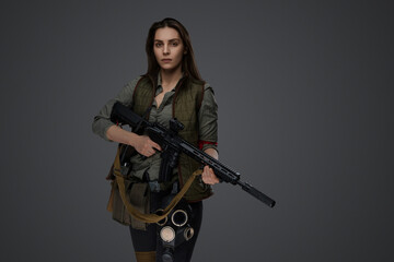 Fototapeta premium Middle Eastern-looking woman dressed in survivalist clothing posing with a rifle against a gray background, portraying strength and resilience
