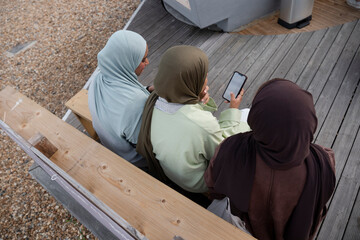 Three Muslim women using smart phone while sitting on bench