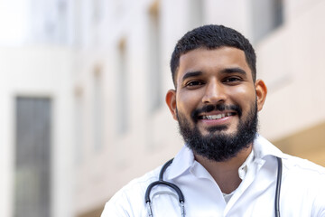 Close-up portrait of a young Indian male doctor standing outside a clinical center in a white coat...