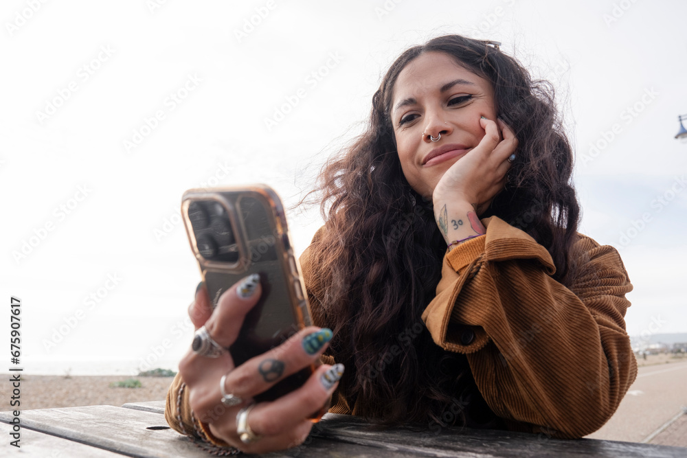 Fototapete smiling woman using smart phone while sitting by picnic table on beach