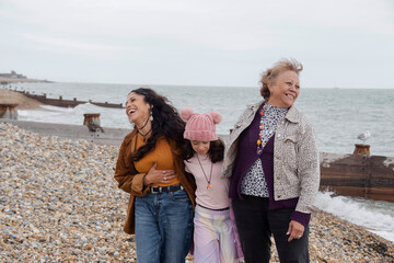 Grandmother, mother and daughter laughing while walking on beach on cloudy day - obrazy, fototapety, plakaty