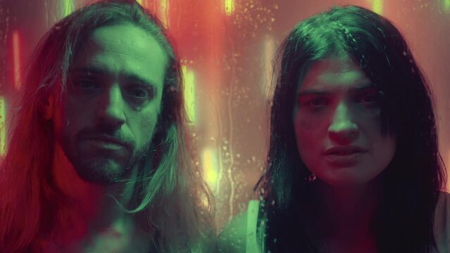 Depressed girl with smudged mascara and sad long-haired man standing in studio with neon light and looking at camera through wet glass. Creative video portrait