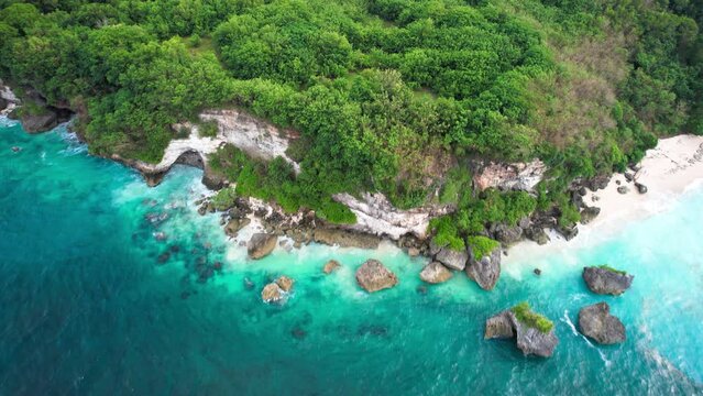 Drone footage flyover green rocky cliffs coastline by turquoise sea and trees in Bali, Indonesia