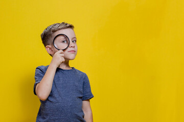 Positive curious schoolboy in casual clothes looking at camera through magnifying glass while standing on yellow background. Copy space - obrazy, fototapety, plakaty