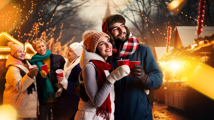 Portrait of young attractive people dressed warm winter outfit enjoying cozy atmosphere outdoor, on Christmas fair.