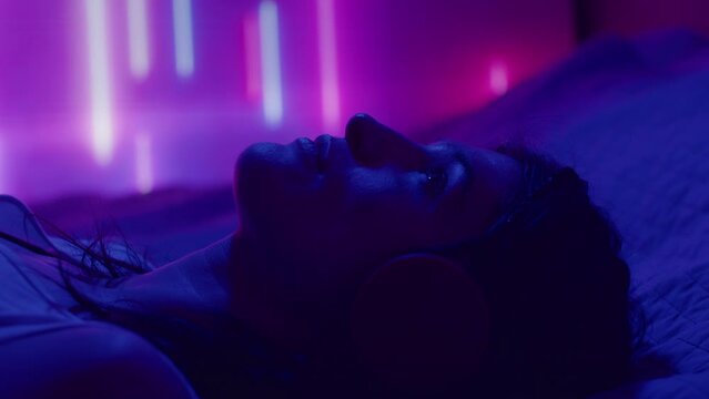 Young Depressed Woman In Headphones Falling Down On Bed, Listening To Music And Staring Up At The Ceiling In Room With Blue And Purple Neon Lights