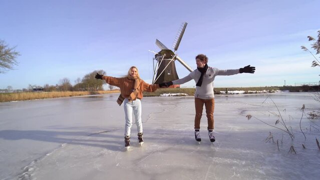 Loving couple having fun on ice in typical dutch landscape with windmill. Woman and man ice skating outdoors in sunny snowy day. Romantic Active date on frozen canal in winter Christmas Eve.