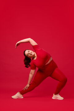 Young overweight woman in sportswear training, doing exercises, stretching against red studio background. Plus size model. Concept of sport, body-positivity, weight loss, body and health care