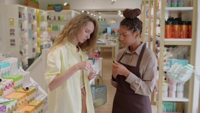 Medium long shot of female consultant helping cheerful customer with choosing face serum for her type of skin at store