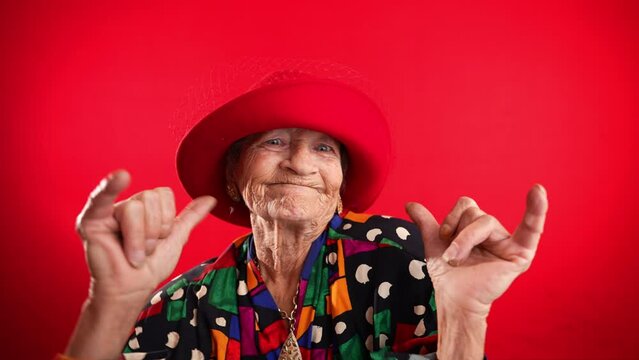 Fisheye portrait of funny elderly mature woman, 80s, wearing red hat giving shaka hand gesture isolated on red background. Concept of old rock and roll person