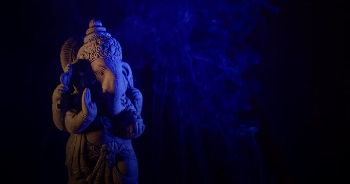 Ganesha idol made of sandstone on a black background at night. There are gradually alternating colorful light colors. There is smoke from the incense that people burn to worship.
