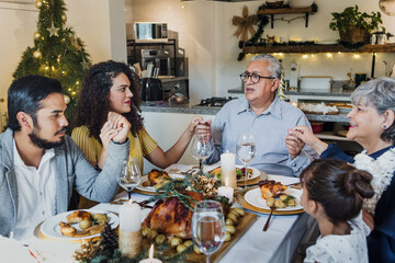 Latin Family praying and holding hands for prayer gathering at thanksgiving dinner at home in...
