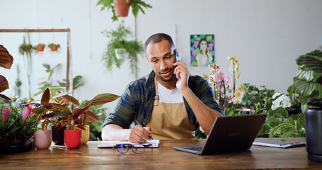 Greenhouse botanist, male botanic worker sitting in own flower shop, typing on laptop and using phone with client. Man florist in flower store orders flowers from greenhouse to be delivered fresh to